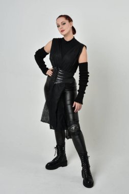 Siyah fütürist bilim kurgu deri kostüm giyen güzel kızıl saçlı kadın modelin tam boy portresi. Dinamik duruş, el hareketleriyle beyaz bir stüdyonun arka planında..