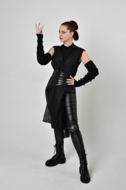 Siyah fütürist bilim kurgu deri kostüm giyen güzel kızıl saçlı kadın modelin tam boy portresi. Dinamik duruş, el hareketleriyle beyaz bir stüdyonun arka planında..
