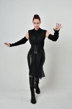 Siyah fütürist bilim kurgu deri kostüm giyen güzel kızıl saçlı kadın modelin tam boy portresi. Dinamik duruş, el hareketleriyle beyaz bir stüdyonun arka planında..