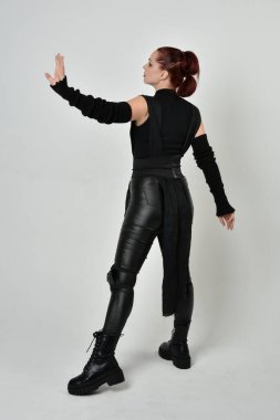 Siyah fütürist bilim kurgu deri kostüm giyen güzel kızıl saçlı kadın modelin tam boy portresi. Dinamik duruş, el hareketleriyle beyaz bir stüdyonun arka planında..