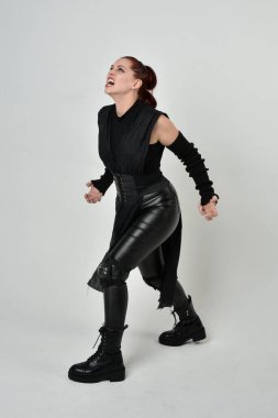 Siyah fütürist bilim kurgu deri kostüm giyen güzel kızıl saçlı kadın modelin tam boy portresi. Dinamik duruş, el hareketleriyle beyaz bir stüdyonun arka planında..