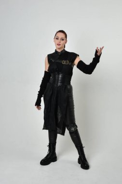 Siyah fütürist bilim kurgu deri kostüm giyen güzel kızıl saçlı kadın modelin tam boy portresi. Dinamik duruş, el hareketleriyle beyaz bir stüdyonun arka planında..