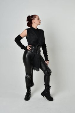 Siyah fütürist bilim kurgu deri kostüm giyen güzel kızıl saçlı kadın modelin tam boy portresi. Dinamik duruş, el hareketleriyle beyaz bir stüdyonun arka planında..