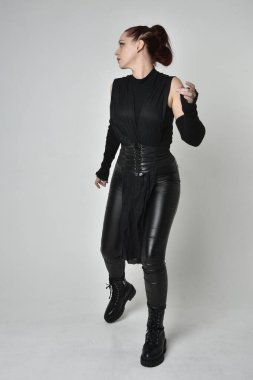 Siyah fütürist bilim kurgu deri kostüm giyen güzel kızıl saçlı kadın modelin tam boy portresi. Dinamik duruş, el hareketleriyle beyaz bir stüdyonun arka planında..