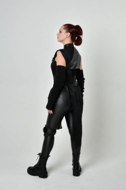 Siyah fütürist bilim kurgu deri kostüm giyen güzel kızıl saçlı kadın modelin tam boy portresi. Dinamik duruş, hareketli eller ile beyaz stüdyo arka planında geriye dönük pozlar.