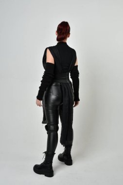 Siyah fütürist bilim kurgu deri kostüm giyen güzel kızıl saçlı kadın modelin tam boy portresi. Dinamik duruş, hareketli eller ile beyaz stüdyo arka planında geriye dönük pozlar.