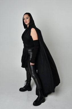 Siyah fütüristik deri kostüm ve siyah pelerinli güzel kızıl saçlı kadın modelin tam boy portresi. Dinamik duruş, hareketli eller ile beyaz stüdyo arka planında geriye dönük pozlar.