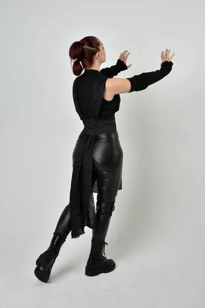 全长肖像漂亮的红头发女模特身穿黑色未来科幻皮衣 动态站立姿势与手势手 面对着背向后从白色工作室背景 — 图库照片