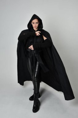 Siyah fütüristik deri kostüm ve siyah pelerinli güzel kızıl saçlı kadın modelin tam boy portresi. Dinamik duruş, hareketli eller ile beyaz stüdyo arka planında geriye dönük pozlar.