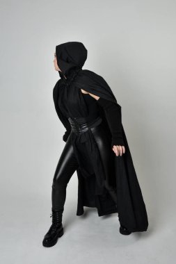 Siyah fütüristik deri kostüm ve siyah pelerinli güzel kızıl saçlı kadın modelin tam boy portresi. Dinamik duruş, hareketli eller ile beyaz stüdyo arka planında geriye dönük pozlar.