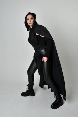 Siyah fütüristik deri kostüm ve siyah pelerinli güzel kızıl saçlı kadın modelin tam boy portresi. Dinamik duruş, hareketli eller ile beyaz stüdyo arka planında geriye dönük pozlar.