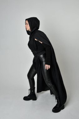 Siyah fütüristik deri kostüm ve siyah pelerinli güzel kızıl saçlı kadın modelin tam boy portresi. Dinamik duruş, hareketli eller ile beyaz stüdyo arka planında geriye dönük pozlar.