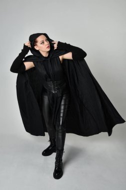 Siyah fütüristik deri kostüm ve siyah pelerinli güzel kızıl saçlı kadın modelin tam boy portresi. Dinamik duruş, hareketli eller ile beyaz stüdyo arka planında geriye dönük pozlar.