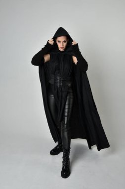 Siyah fütüristik deri kostüm ve siyah pelerinli güzel kızıl saçlı kadın modelin tam boy portresi. Dinamik duruş, hareketli eller ile beyaz stüdyo arka planında geriye dönük pozlar.