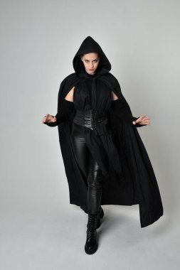 Siyah fütüristik deri kostüm ve siyah pelerinli güzel kızıl saçlı kadın modelin tam boy portresi. Dinamik duruş, hareketli eller ile beyaz stüdyo arka planında geriye dönük pozlar.