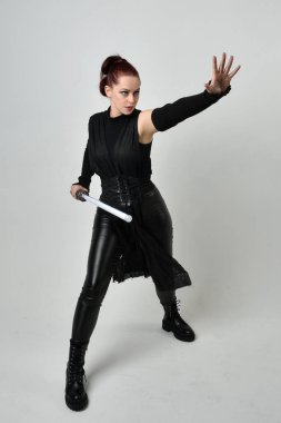 Siyah fütüristik deri kostüm giyen, elinde ışın kılıcı olan kızıl saçlı bir kadın modelin tam boy portresi. Dinamik duruş, hareketli eller ile beyaz stüdyo arka planında geriye dönük pozlar.