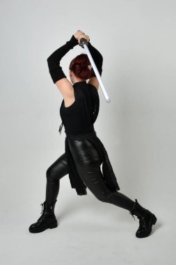 Siyah fütüristik deri kostüm giyen, elinde ışın kılıcı olan kızıl saçlı bir kadın modelin tam boy portresi. Dinamik duruş, hareketli eller ile beyaz stüdyo arka planında geriye dönük pozlar.