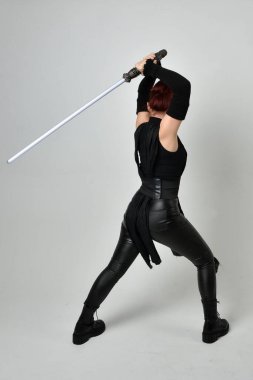 Full length portrait of pretty red haired female model wearing black futuristic scifi leather costume, holding a lightsaber sword weapon. Dynamic standing poses with gestural hands, facing backwards away from  on a white studio background. clipart