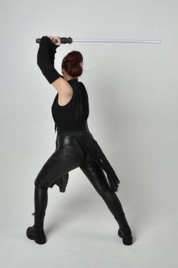 Siyah fütüristik deri kostüm giyen, elinde ışın kılıcı olan kızıl saçlı bir kadın modelin tam boy portresi. Dinamik duruş, hareketli eller ile beyaz stüdyo arka planında geriye dönük pozlar.