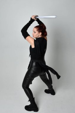 Siyah fütüristik deri kostüm giyen, elinde ışın kılıcı olan kızıl saçlı bir kadın modelin tam boy portresi. Dinamik duruş, hareketli eller ile beyaz stüdyo arka planında geriye dönük pozlar.