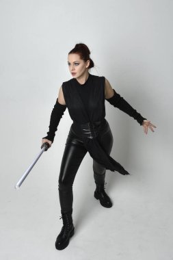 Siyah fütüristik deri kostüm giyen, elinde ışın kılıcı olan kızıl saçlı bir kadın modelin tam boy portresi. Dinamik duruş, hareketli eller ile beyaz stüdyo arka planında geriye dönük pozlar.