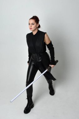 Siyah fütüristik deri kostüm giyen, elinde ışın kılıcı olan kızıl saçlı bir kadın modelin tam boy portresi. Dinamik duruş, hareketli eller ile beyaz stüdyo arka planında geriye dönük pozlar.