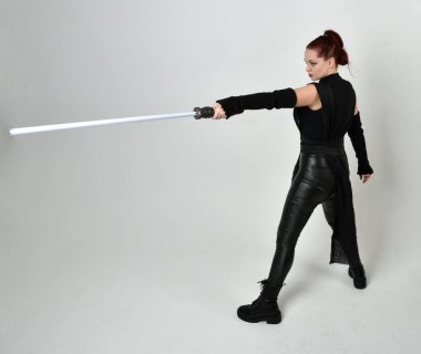 Siyah fütüristik deri kostüm giyen, elinde ışın kılıcı olan kızıl saçlı bir kadın modelin tam boy portresi. Dinamik duruş, hareketli eller ile beyaz stüdyo arka planında geriye dönük pozlar.