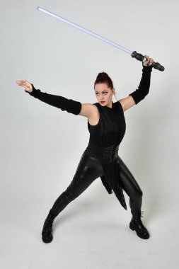 Siyah fütüristik deri kostüm giyen, elinde ışın kılıcı olan kızıl saçlı bir kadın modelin tam boy portresi. Dinamik duruş, hareketli eller ile beyaz stüdyo arka planında geriye dönük pozlar.