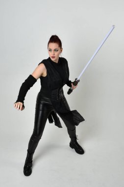 Siyah fütüristik deri kostüm giyen, elinde ışın kılıcı olan kızıl saçlı bir kadın modelin tam boy portresi. Dinamik duruş, hareketli eller ile beyaz stüdyo arka planında geriye dönük pozlar.