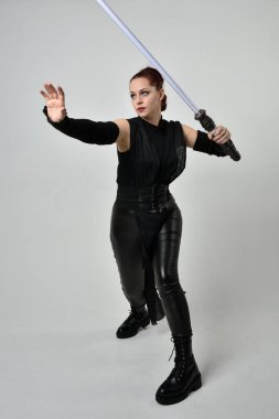 Siyah fütüristik deri kostüm giyen, elinde ışın kılıcı olan kızıl saçlı bir kadın modelin tam boy portresi. Dinamik duruş, hareketli eller ile beyaz stüdyo arka planında geriye dönük pozlar.