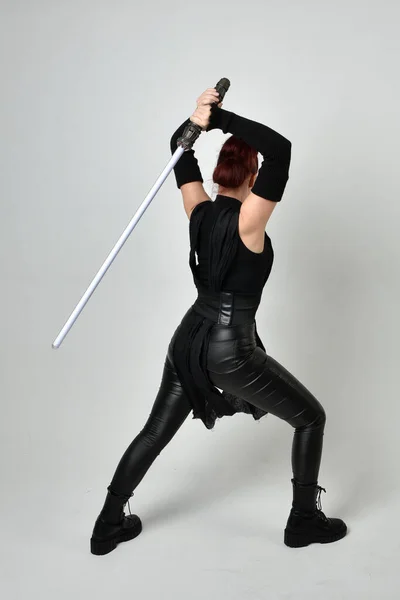 黒の未来的なScifi革の衣装を身に着けているかなり赤い髪の女性モデルの完全な長さの肖像画は ライトセーバーの剣の武器を保持 白いスタジオの背景から後ろに向かって ジェスチャーの手でダイナミックな立ち位置 — ストック写真
