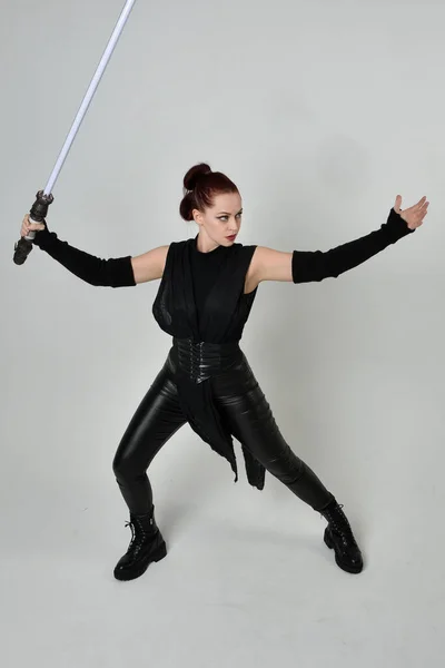 全长肖像漂亮的红头发女模特身穿黑色未来科幻皮衣 手持光剑武器 动态站立姿势与手势手 面对着背向后从白色工作室背景 — 图库照片