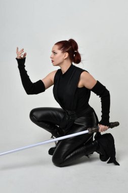 Siyah fütüristik deri kostüm giyen, elinde ışın kılıcı olan kızıl saçlı bir kadın modelin tam boy portresi. Dinamik oturma pozu beyaz stüdyo arka planında.