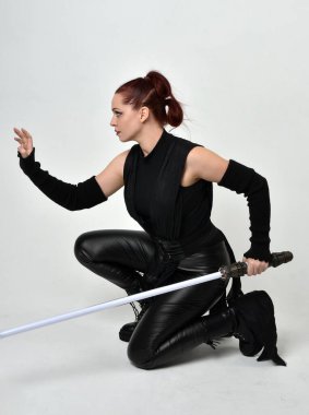 Siyah fütüristik deri kostüm giyen, elinde ışın kılıcı olan kızıl saçlı bir kadın modelin tam boy portresi. Dinamik oturma pozu beyaz stüdyo arka planında.