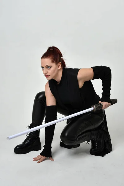全长肖像漂亮的红头发女模特身穿黑色未来科幻皮衣 手持光剑武器 在白色的工作室背景上动态地坐姿 — 图库照片