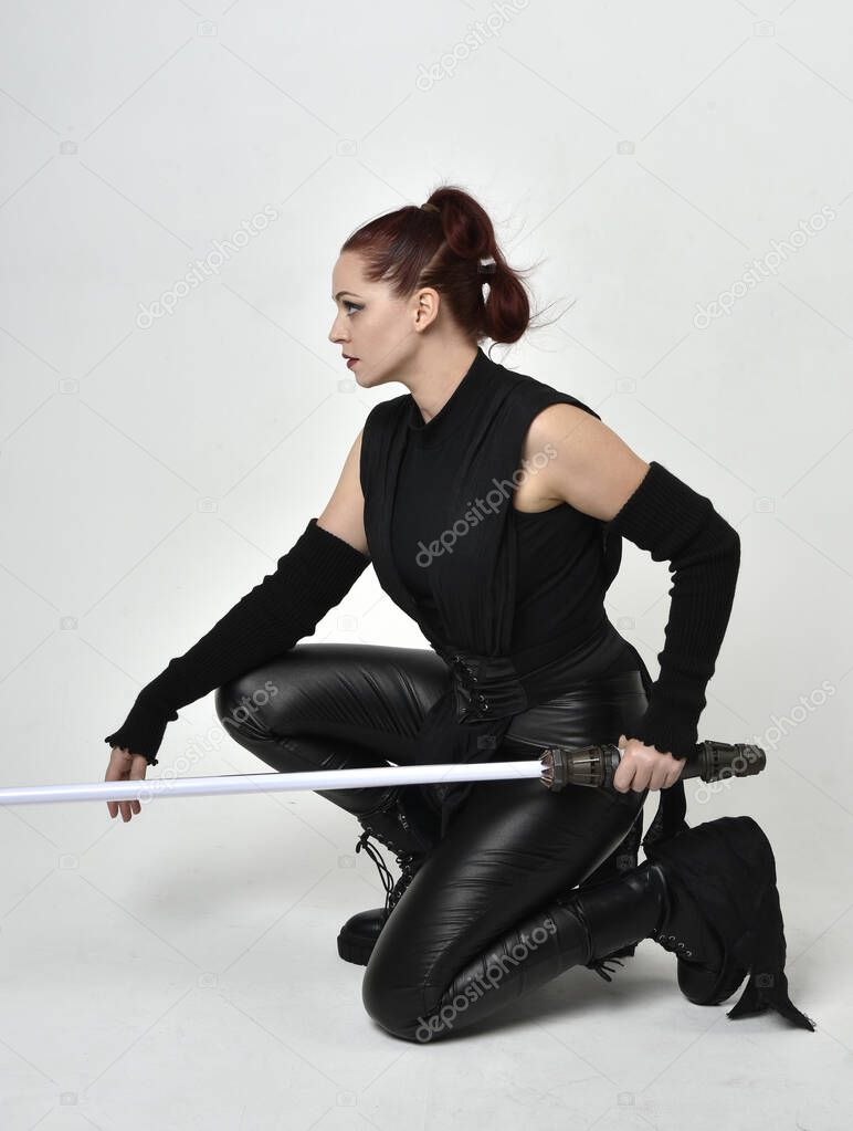 Full length portrait of pretty red haired female model wearing black futuristic scifi leather costume, holding a lightsaber sword weapon. Dynamic sitting pose on a white studio background.