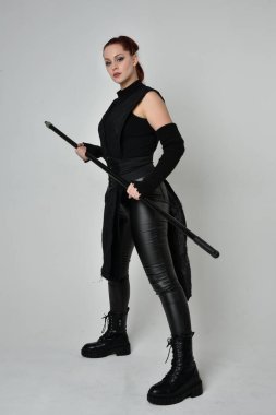 Uzun boylu, kızıl saçlı, siyah fütürist deri kostüm giyen, elinde mızrak tutan bir kadın modelin portresi. Beyaz stüdyo arka planında dinamik duruş.