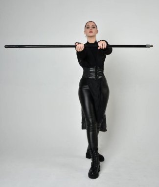 Uzun boylu, kızıl saçlı, siyah fütürist deri kostüm giyen, elinde mızrak tutan bir kadın modelin portresi. Beyaz stüdyo arka planında dinamik duruş.