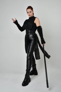 Uzun boylu, kızıl saçlı, siyah fütürist deri kostüm giyen, elinde mızrak tutan bir kadın modelin portresi. Beyaz stüdyo arka planında dinamik duruş.