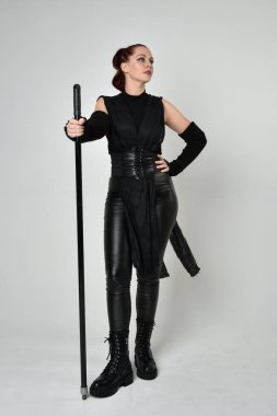 Uzun boylu, kızıl saçlı, siyah fütürist deri kostüm giyen, elinde mızrak tutan bir kadın modelin portresi. Beyaz stüdyo arka planında dinamik duruş.