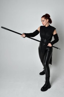 Uzun boylu, kızıl saçlı, siyah fütürist deri kostüm giyen, elinde mızrak tutan bir kadın modelin portresi. Beyaz stüdyo arka planında dinamik duruş.