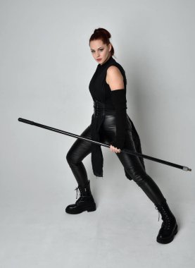 Uzun boylu, kızıl saçlı, siyah fütürist deri kostüm giyen, elinde mızrak tutan bir kadın modelin portresi. Beyaz stüdyo arka planında dinamik duruş.