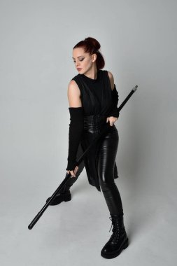 Uzun boylu, kızıl saçlı, siyah fütürist deri kostüm giyen, elinde mızrak tutan bir kadın modelin portresi. Beyaz stüdyo arka planında dinamik duruş.