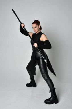 Uzun boylu, kızıl saçlı, siyah fütürist deri kostüm giyen, elinde mızrak tutan bir kadın modelin portresi. Beyaz stüdyo arka planında dinamik duruş.