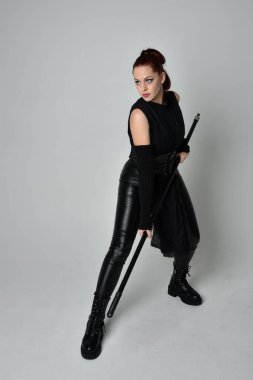 Uzun boylu, kızıl saçlı, siyah fütürist deri kostüm giyen, elinde mızrak tutan bir kadın modelin portresi. Beyaz stüdyo arka planında dinamik duruş.