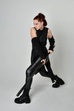Uzun boylu, kızıl saçlı, siyah fütürist deri kostüm giyen, elinde mızrak tutan bir kadın modelin portresi. Beyaz stüdyo arka planında dinamik duruş.