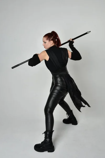 全长肖像漂亮的红头发女模特身穿黑色未来科幻皮衣 手持长矛武器 白色工作室背景上的动态站立姿势 — 图库照片