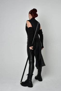 Siyah fütüristik deri kostüm giyen ve elinde mızrak tutan kızıl saçlı güzel bir kadın modelin tam boy portresi. Dinamik duruş pozisyonu ters, beyaz stüdyo arka planı.