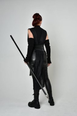 Siyah fütüristik deri kostüm giyen ve elinde mızrak tutan kızıl saçlı güzel bir kadın modelin tam boy portresi. Dinamik duruş pozisyonu ters, beyaz stüdyo arka planı.