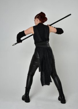 Siyah fütüristik deri kostüm giyen ve elinde mızrak tutan kızıl saçlı güzel bir kadın modelin tam boy portresi. Dinamik duruş pozisyonu ters, beyaz stüdyo arka planı.